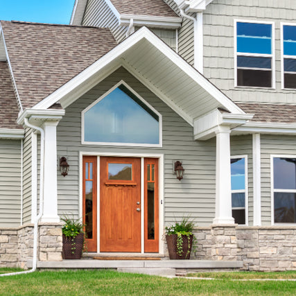 DoorsA modern suburban home with a gable roof, gray siding, and stone accents. A wooden front door is flanked by potted plants, conveying a welcoming tone.
