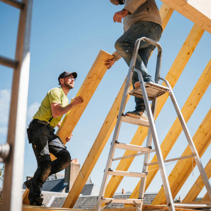 Building MaterialsTwo construction workers assemble a wooden roof frame. One stands on a ladder, the other hands him a beam. The scene conveys teamwork under a clear blue sky.